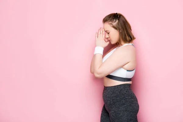 Vista laterale della ragazza in sovrappeso in abbigliamento sportivo in piedi con le mani in preghiera sul rosa — Foto stock