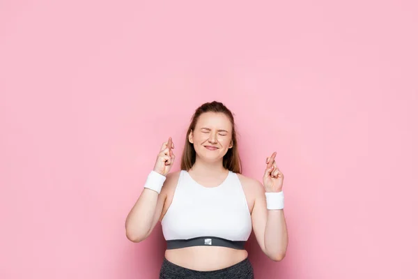 Chica con sobrepeso cierra los ojos sosteniendo los dedos cruzados en rosa - foto de stock