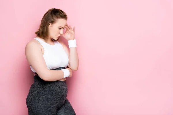 Thoughtful overweight girl in sportswear touching head on pink — Stock Photo