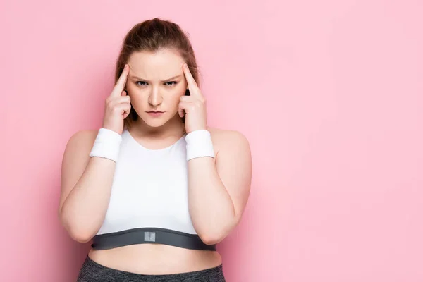 Seria, chica con sobrepeso reflexivo tocando la cabeza mientras mira a la cámara en rosa - foto de stock