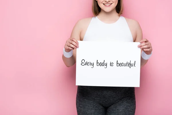 Vista cortada de sorrir menina com excesso de peso segurando cartaz com cada corpo é bela inscrição em rosa — Fotografia de Stock