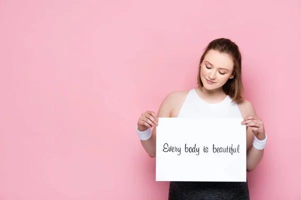 Smiling overweight girl holding placard with every body is beautiful inscription on pink — Stock Photo