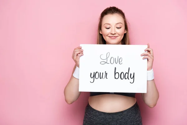 Happy overweight girl with closed eyes holding placard with love your body lettering on pink — Stock Photo