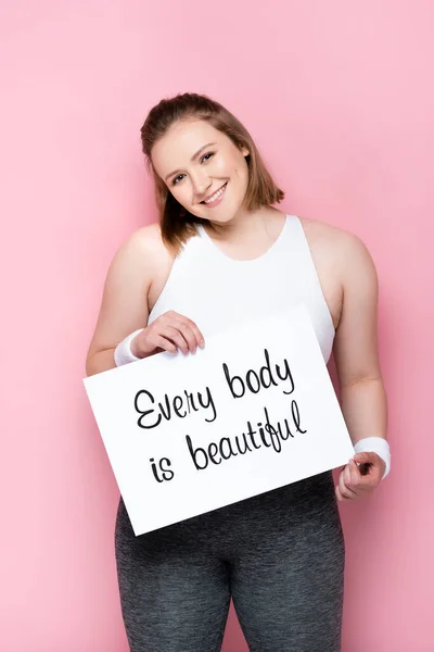 Cheerful overweight girl holding placard with every body is beautiful inscription on pink — Stock Photo