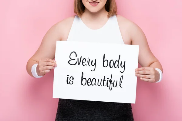 Cropped view of smiling overweight girl holding placard with every body is beautiful inscription on pink — Stock Photo