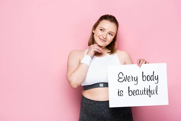 Smiling overweight girl holding placard with every body is beautiful inscription on pink — Stock Photo