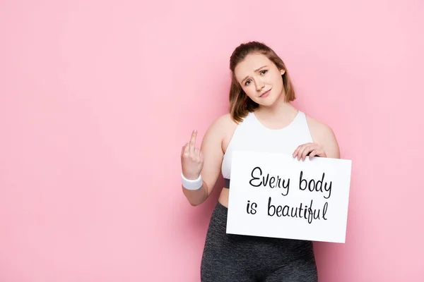 Offended overweight girl showing middle finger while holding placard with every body is beautiful lettering on pink — Stock Photo