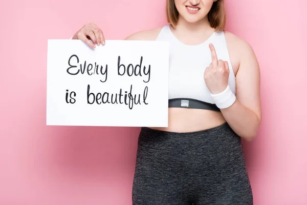 Cropped view of overweight girl showing middle finger while holding placard with every body is beautiful inscription on pink — Stock Photo