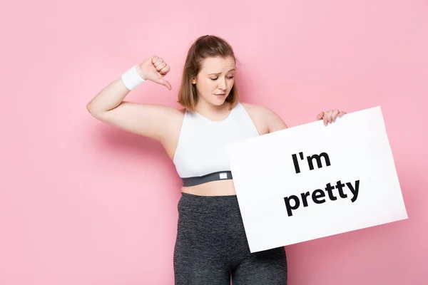 Ofendido chica con sobrepeso sosteniendo pancarta con estoy bastante inscripción y mostrando el pulgar hacia abajo en rosa - foto de stock