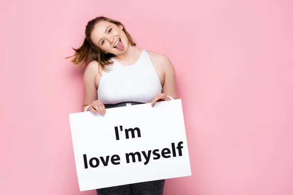 Cheerful overweight girl holding placard with i love myself inscription and sticking out tongue on pink — Stock Photo