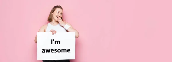 Flirty overweight girl holding placard with i am awesome lettering and covering mouth with hand isolated on pink, panoramic shot — Stock Photo