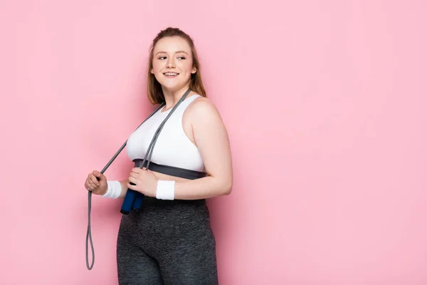 Smiling overweight girl with jumping rope around neck looking away on pink — Stock Photo