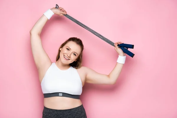 Cheerful overweight girl holding jumping rope above head while looking at camera on pink — Stock Photo