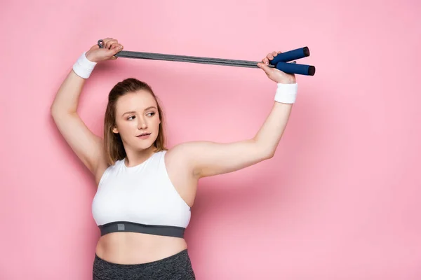 Pretty plus size girl holding jumping rope above head while looking away on pink — Stock Photo
