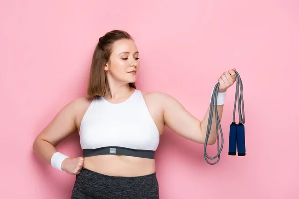 Confident plus size girl holding jumping rope while standing with hand on hip on pink — Stock Photo