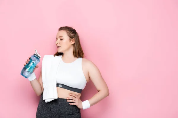 Confident overweight girl with towel on shoulder holding sports bottle while standing with hand on hip on pink — Stock Photo