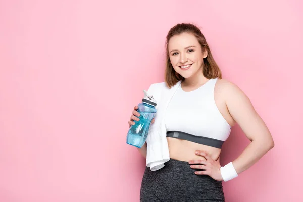 Sourire plus la taille fille avec serviette sur l'épaule tenant bouteille de sport tout en se tenant avec la main sur la hanche sur rose — Photo de stock