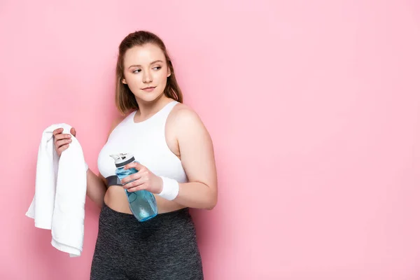 Confiado chica con sobrepeso sosteniendo botella de deportes y toalla blanca mientras mira hacia otro lado en rosa - foto de stock