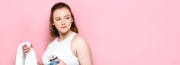 Pretty plus size girl holding sports bottle and white towel while looking away on pink — Stock Photo