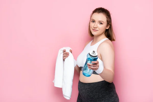 Sonriente chica con sobrepeso sosteniendo botella deportiva y toalla blanca en rosa - foto de stock