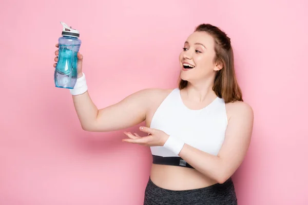 Excitada chica con sobrepeso sosteniendo botella deportiva en la mano extendida en rosa - foto de stock