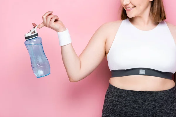 Vue recadrée de sourire fille en surpoids tenant bouteille de sport sur rose — Photo de stock