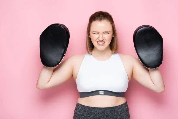 Angry overweight girl in boxing pads grimacing while looking at camera on pink — Stock Photo