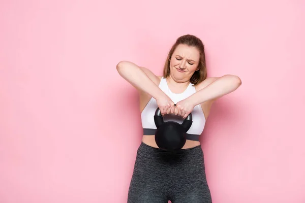 Tamanho mais menina em sportswear grimacing ao levantar peso pesado em rosa — Fotografia de Stock