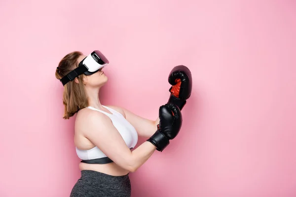 Vista lateral de la chica con sobrepeso en auriculares vr y guantes de boxeo mirando hacia arriba en rosa - foto de stock