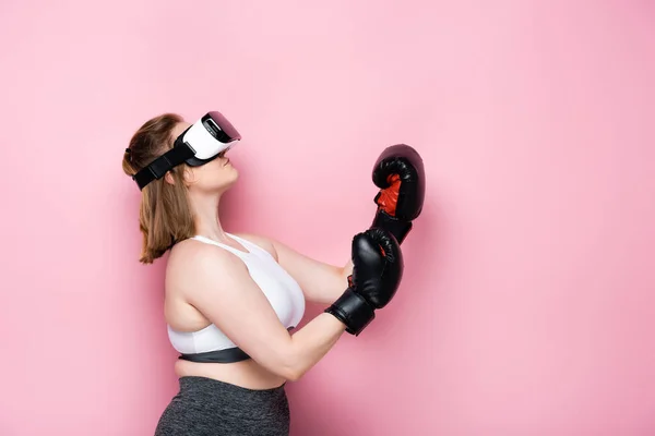 Vista laterale della ragazza in sovrappeso in auricolare vr e guanti da boxe guardando in alto sul rosa — Foto stock