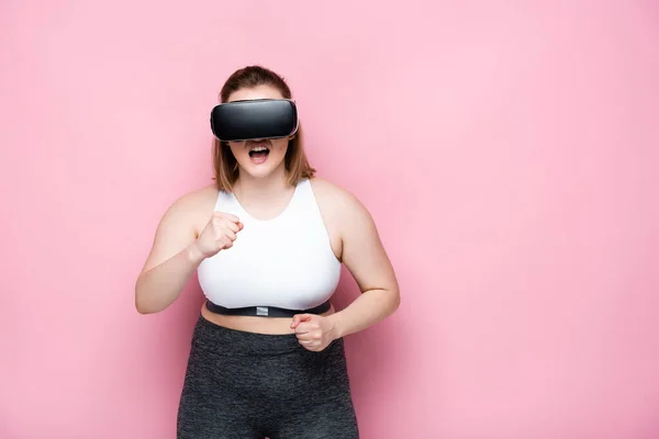 Chica enojada con sobrepeso en ropa deportiva y auriculares vr con puños cerrados en rosa - foto de stock