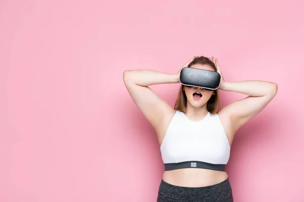 Shocked size plus girl in sportswear and vr headset touching head on pink — стоковое фото