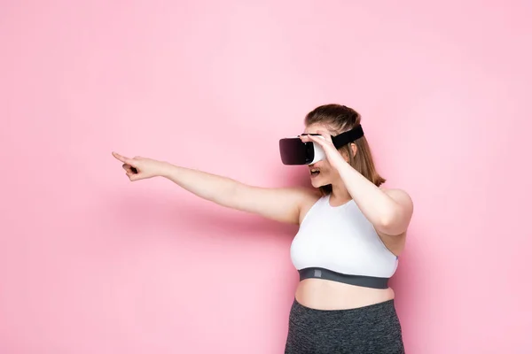 Chica con sobrepeso en ropa deportiva y auriculares vr apuntando con el dedo en rosa - foto de stock