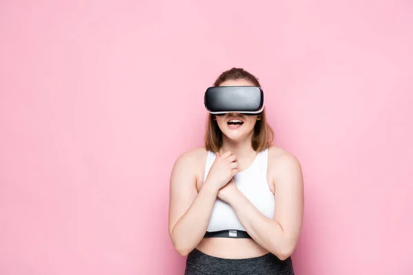 Excited size plus girl in sportswear and vr headset holding hands on chest on pink — Stock Photo