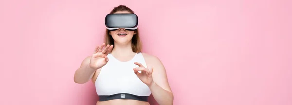 Colpo panoramico di dimensioni sorridenti più ragazza in auricolare vr e abbigliamento sportivo sorridente e gestuale su rosa — Foto stock