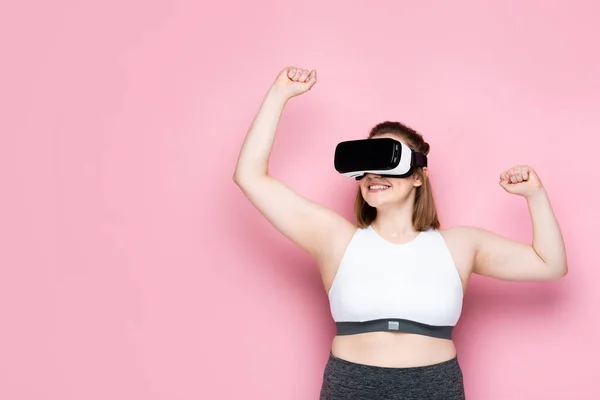 Dimensione eccitato più ragazza in abbigliamento sportivo e auricolare vr mostrando gesto vincitore sul rosa — Foto stock