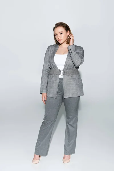 Full length view of elegant, beautiful overweight girl touching hair while posing on grey — Stock Photo