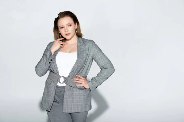 Stylish overweight girl touching face while standing with hand on hip on grey — Stock Photo