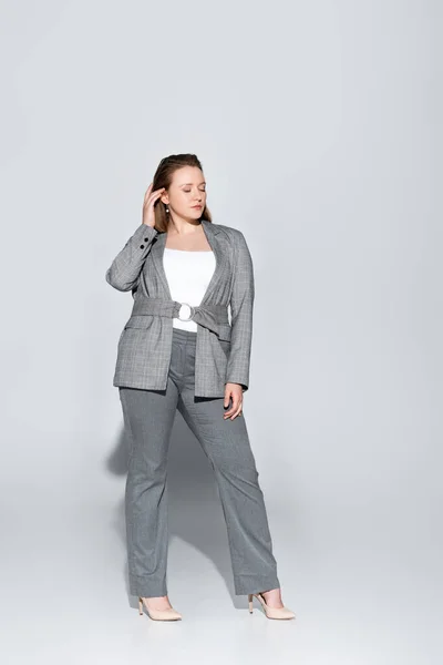 Full length view of stylish overweight girl touching hair while posing with closed eyes on grey — Stock Photo