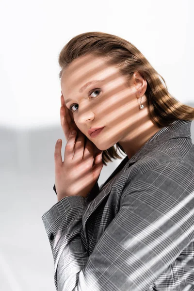 Bella ragazza in sovrappeso che si tiene per mano vicino al viso mentre guarda la fotocamera su bianco alla luce del sole con ombre — Foto stock