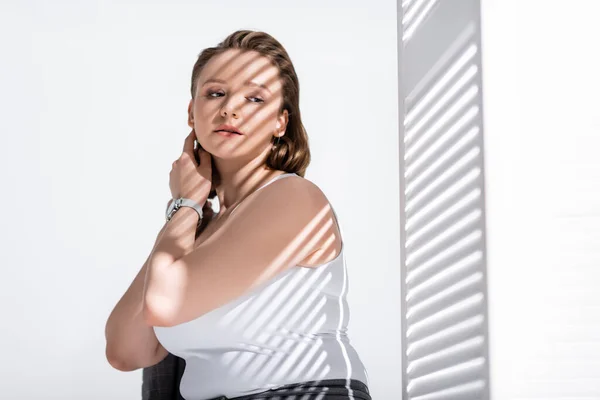 Beautiful, dreamy overweight girl in white top touching neck while posing on white in sunlight with shadows — Stock Photo