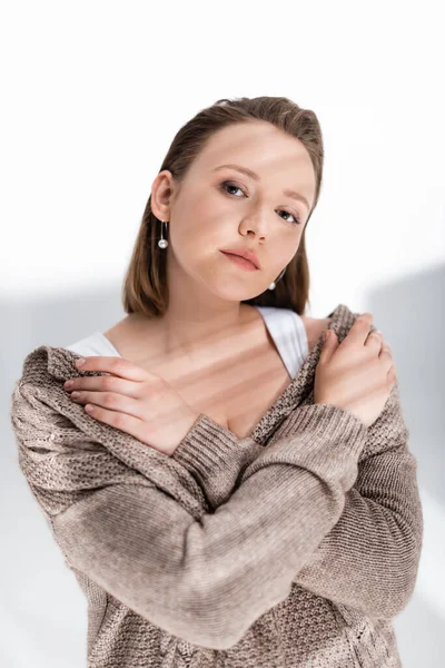 Красивая, уверенная в себе толстая девушка в сером свитере обнимает себя и смотрит на камеру на белом с солнечным светом и тенями — стоковое фото