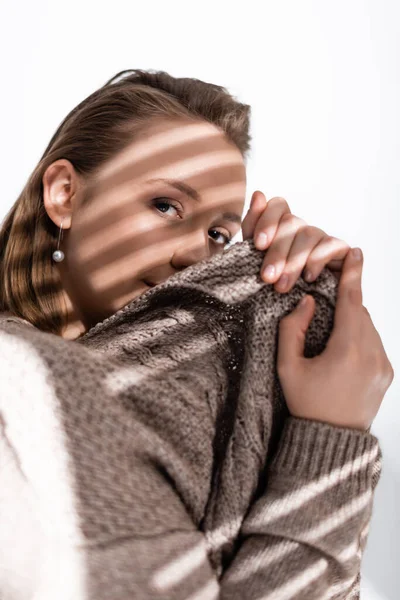 Attraente ragazza in sovrappeso oscurando il viso con maglione grigio e guardando la fotocamera su bianco con luce solare e ombre — Foto stock
