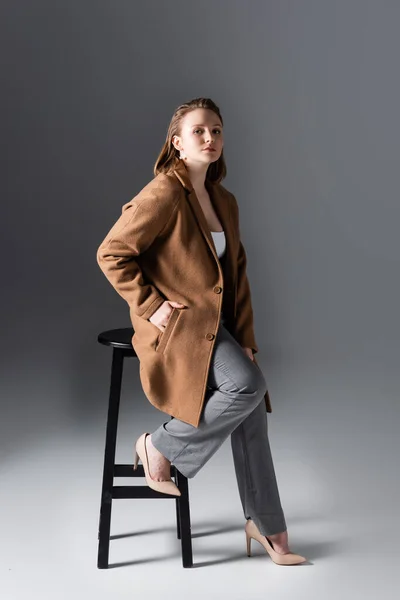 Full length view of serious, attractive overweight girl posing on high stool with hand in pocket on grey — Stock Photo