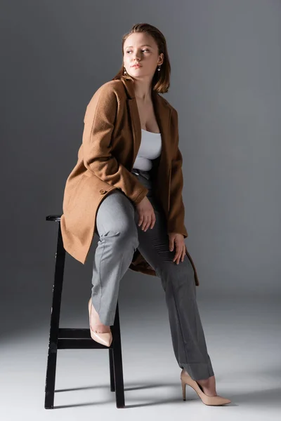 Full length view of confident, stylish overweight girl posing on high chair and looking away on grey — Stock Photo