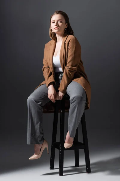 Full length view of confident, pretty overweight girl sitting on high chair and looking at camera while posing on grey — Stock Photo