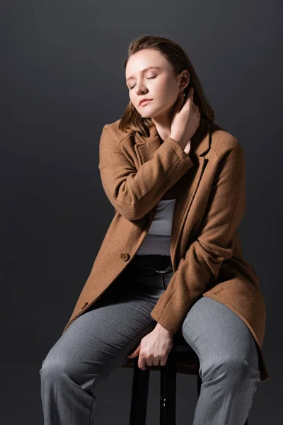 Menina bonita plus size sentada na cadeira e tocando o pescoço enquanto posando com os olhos fechados isolados no cinza — Fotografia de Stock