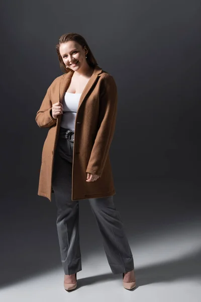 Full length view of smiling, beautiful plus size girl in trendy clothing looking at camera on grey — Stock Photo