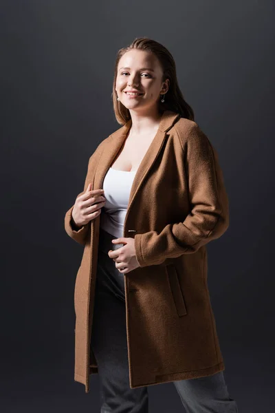 Улыбающаяся, привлекательная толстая девушка в пальто, смотрящая на камеру, изолированную на сером — стоковое фото