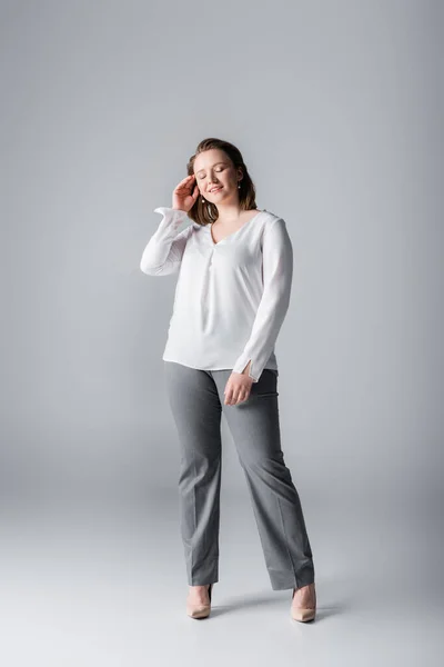 Piena lunghezza vista di elegante ragazza in sovrappeso toccare i capelli mentre posa con gli occhi chiusi sul grigio — Foto stock